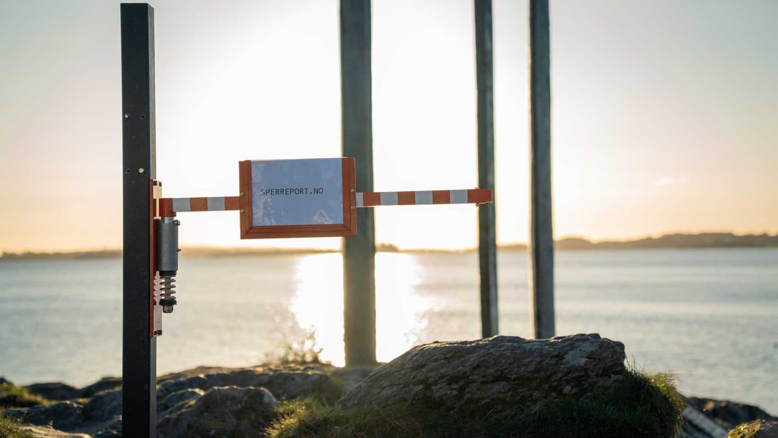 Sperreport med fjærhengsel og skiltholder i motlys, med solnedgangen speilet i fjorden bak