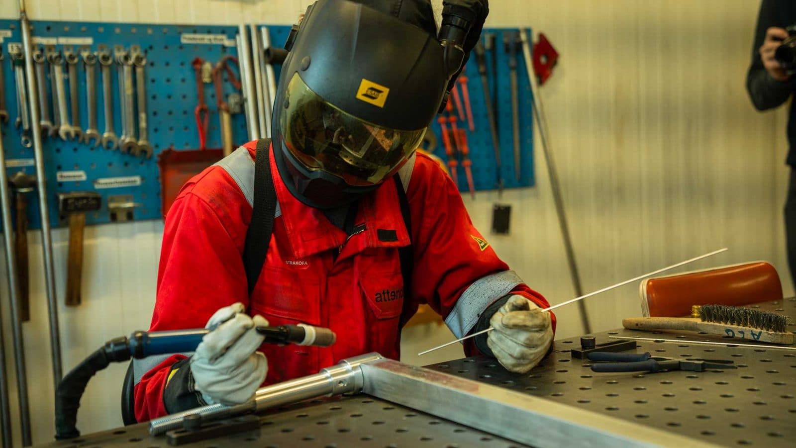 Sveiser i rødt vernetøy med sveisemaske arbeider med en aluminiumsprofil på sveisebordet, verktøytavle i bakgrunnen