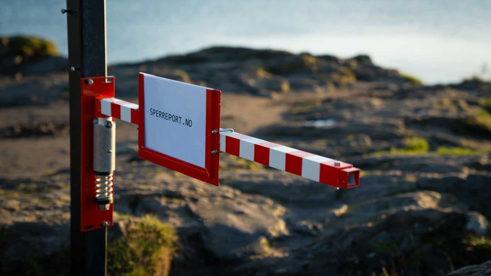 Nærbilde av spiralfjæren og skiltholderen på en sperreport montert på svart stolpe, med svaberg i bakgrunnen