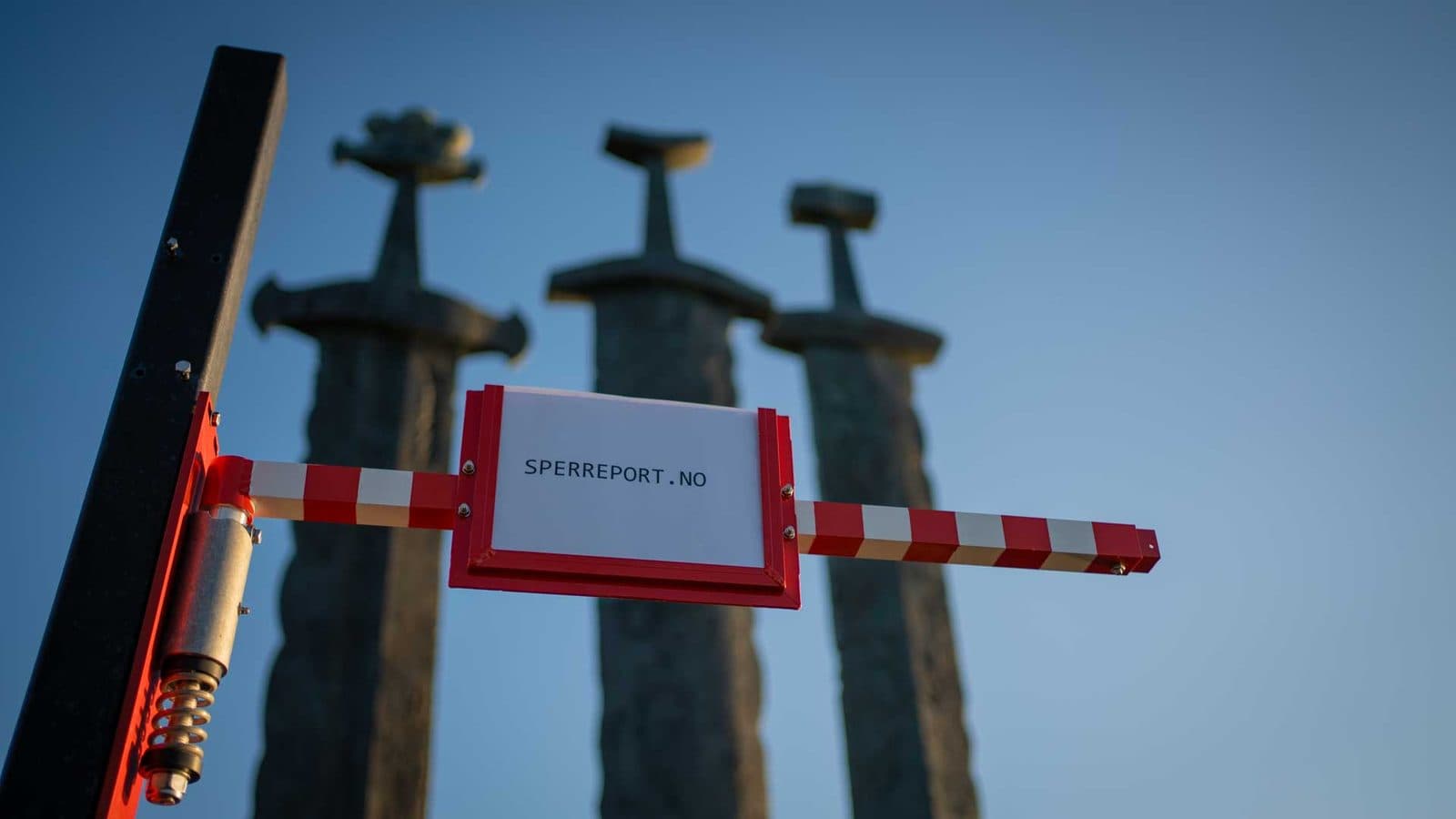 Nærbilde av det fjærbelastede hengselet på en sperreport sideveis — rød brakett og spiralfjær på stolpen, med rød/hvit-stripet arm og skiltholder foran Sverd i fjell