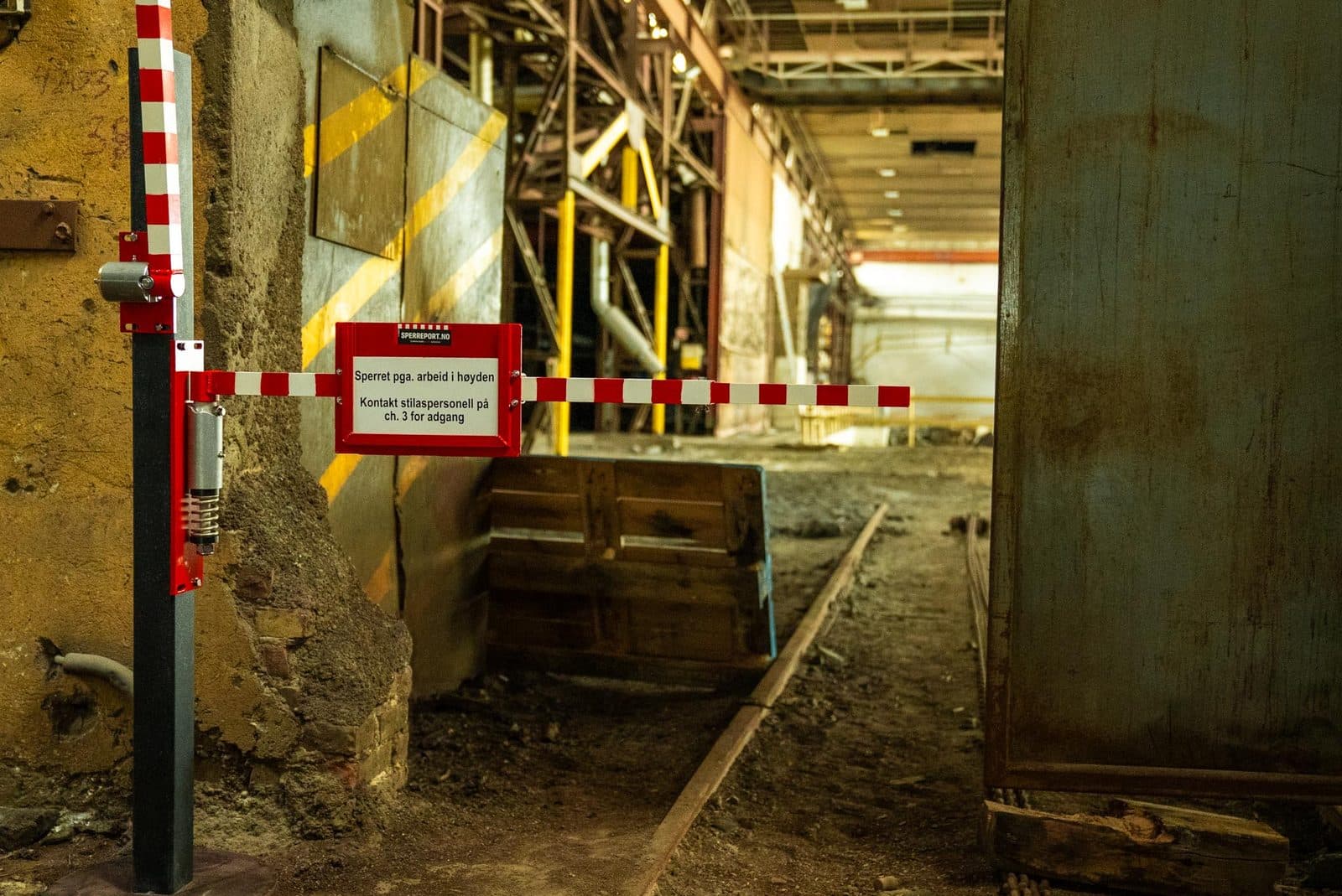 Sperreport vertikal med armen felt opp langs stolpen i en industrihall, med skiltholder og rødt HMS-skilt om arbeid i høyden