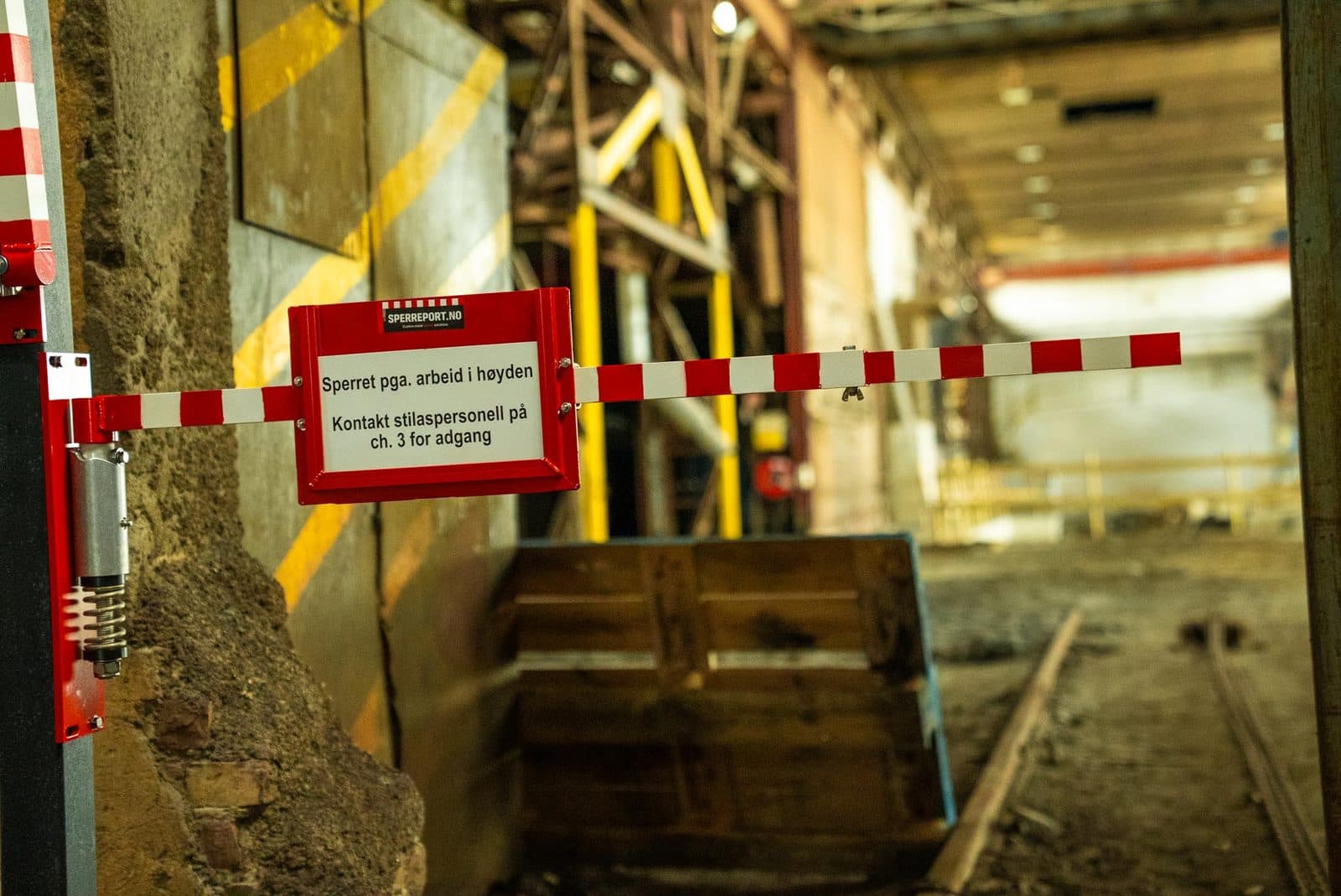 Nærbilde av hengselet og fjærmekanismen på en sperreport montert mot betongvegg i industrihall